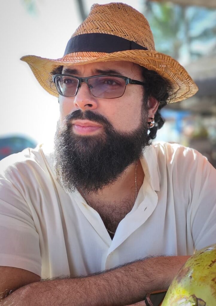 Na foto - Moisés Tedeschi usando chapéu de palha, na praia (em um dia de sol), de óculos e olhando para a direita.